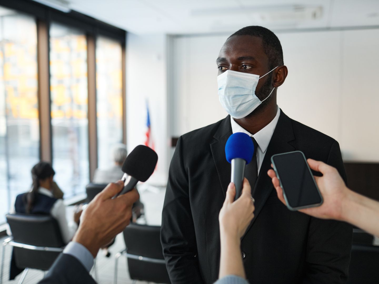 African American businessman in formal attire speaks to media while wearing a face mask in a conference room setting.